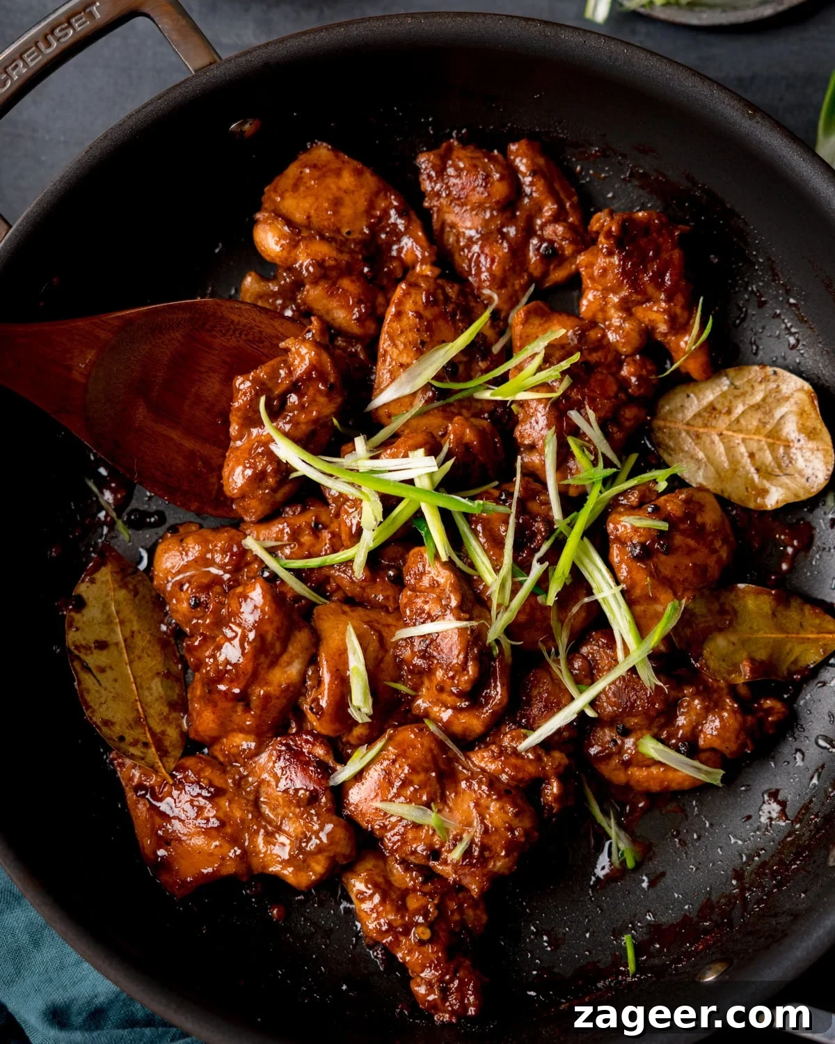 Authentic Filipino Chicken Adobo 2 Overhead image of chicken adobo in a dark glaze with bay leaves, topped with sliced spring onions, served in a black frying pan with a wooden spoon. A Filipino Chicken dish made with pan fried chicken thigh fillets in a tangy, garlicky sauce.