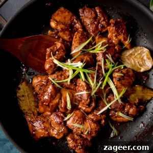 Chicken adobo in a dark glaze with bay leaves, topped with sliced spring onions, served in a black frying pan with a wooden spoon, ready to be served over rice.