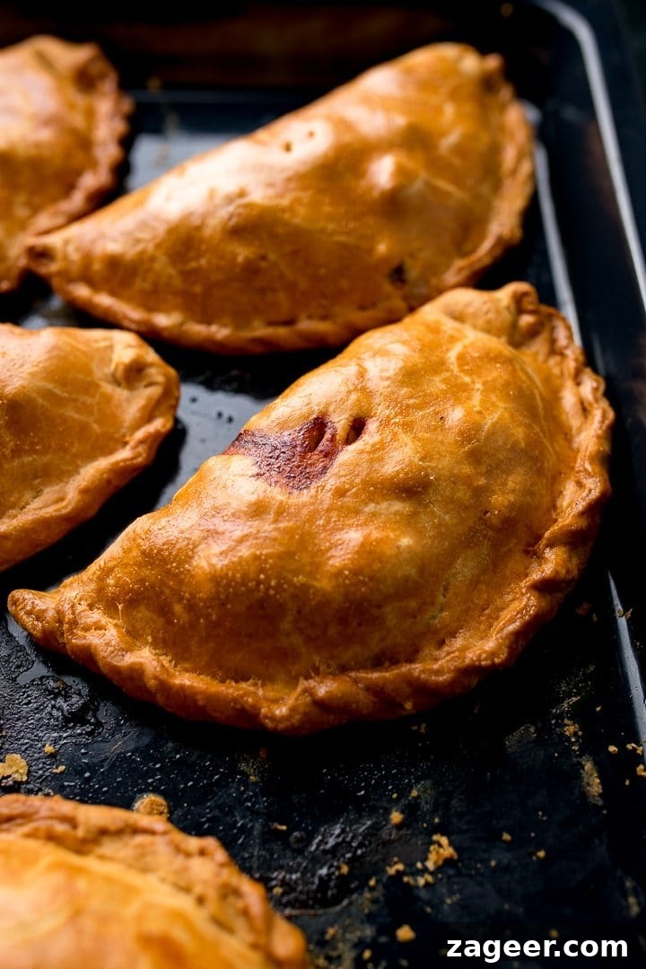 Authentic Cornish Pasty Recipe: Traditional British Meat Pasty Guide 2 Golden brown Cornish Pasties are arranged on a dark baking tray, freshly baked with a flaky pastry.
