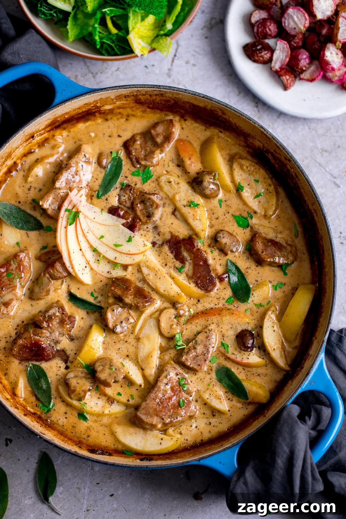 Creamy Pork and Apple Casserole Recipe with Herbed Crust 6 Overhead image of creamy pork and apple casserole in a blue casserole dish on a light background. Side dishes of cabbage and radishes are just in shot.