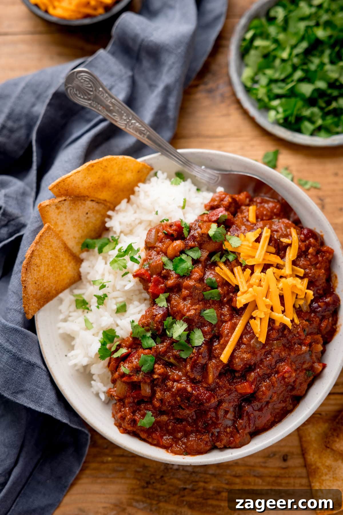 Authentic Chilli Con Carne Recipe with Smoky Beef and Beans 2 A bowl of chilli con carne with kidney beans, minced beef, cheese, and coriander over white rice. Tortilla chips on the side and a blue napkin to the side of the bowl.