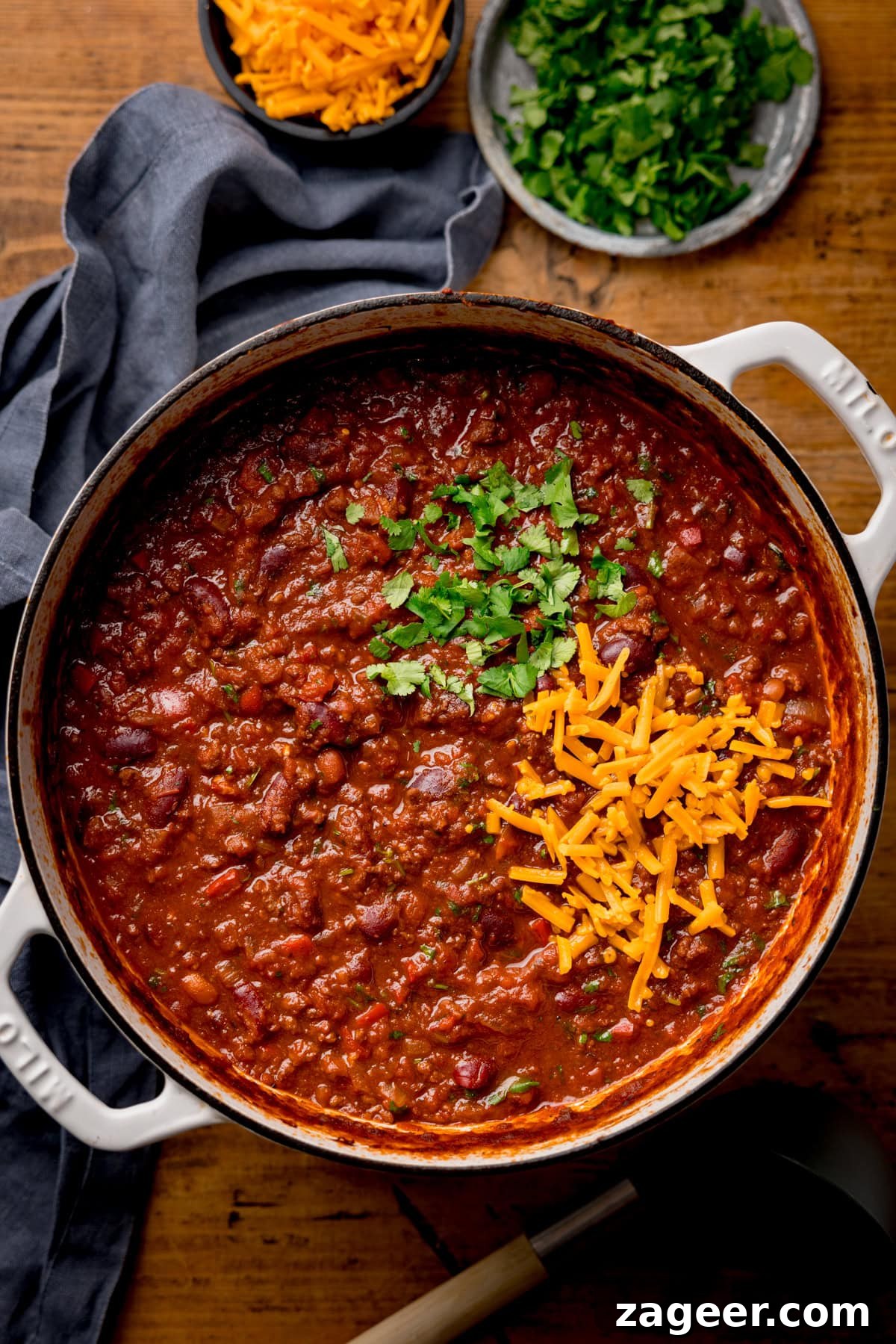 Authentic Chilli Con Carne Recipe with Smoky Beef and Beans 6 A pot of hearty chilli con carne topped with fresh coriander and grated cheddar, beside small bowls of coriander and grated cheese on a wooden table.