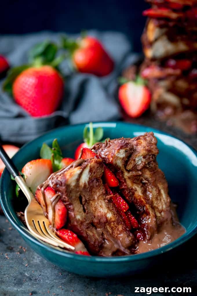 Slice of chocolate bread and butter pudding cake filled and topped with Nutella and strawberries in a dark green bowl with a gold fork with black handle. Scattered strawberries and navy blue linen napkin in the background.