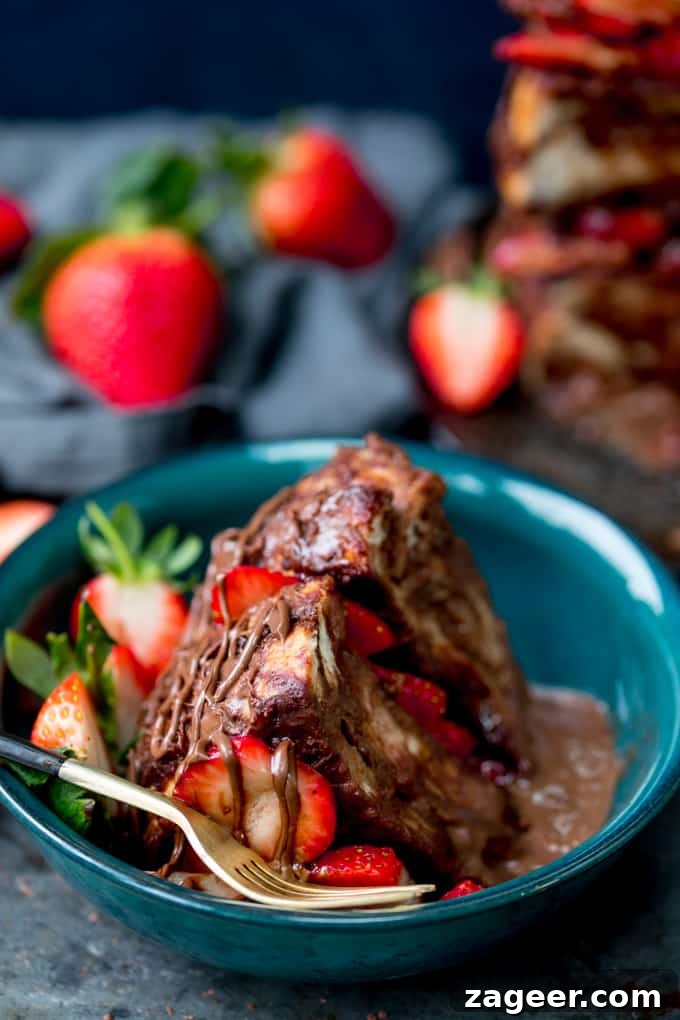 Slice of chocolate bread and butter pudding cake filled and topped with Nutella and strawberries in a dark green bowl with a gold fork with black handle. Drizzled with Nutella. Scattered strawberries and navy blue linen napkin in the background.