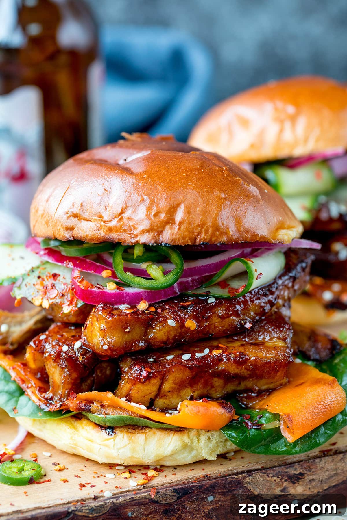 Sticky Glazed Pork Burger with Quick Pickled Veggies 2 Side on image of a sticky pork belly burger with carrot, spinach, red onion and jalapeños on a brioche bun. The burger is on a wooden board and there is a further burger in the background, just in shot.
