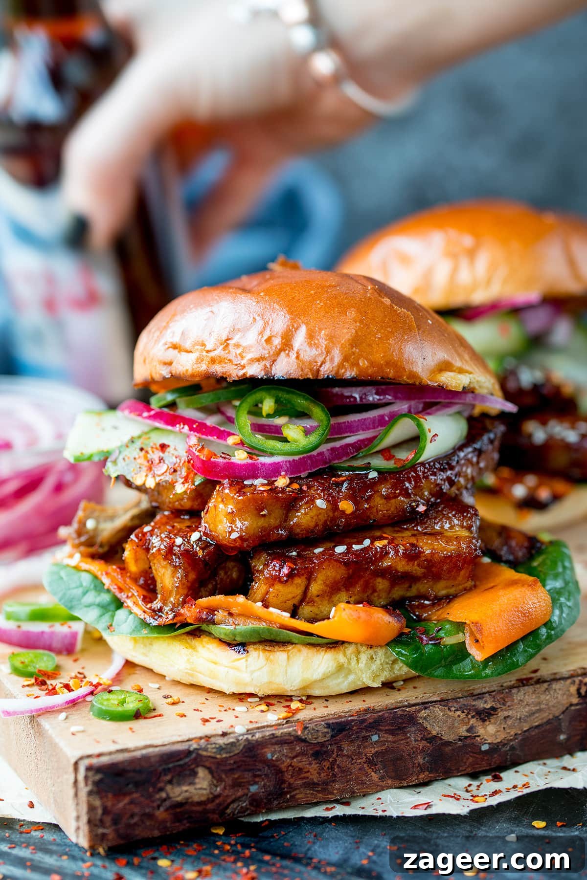 Sticky Glazed Pork Burger with Quick Pickled Veggies 5 Side on image of a sticky pork belly burger with carrot, spinach, red onion and jalapeños on a brioche bun. The burger is on a wooden board and there is a further burger in the background, just in shot. There is also a hand picking up a bottle of beer in the rear of the shot.