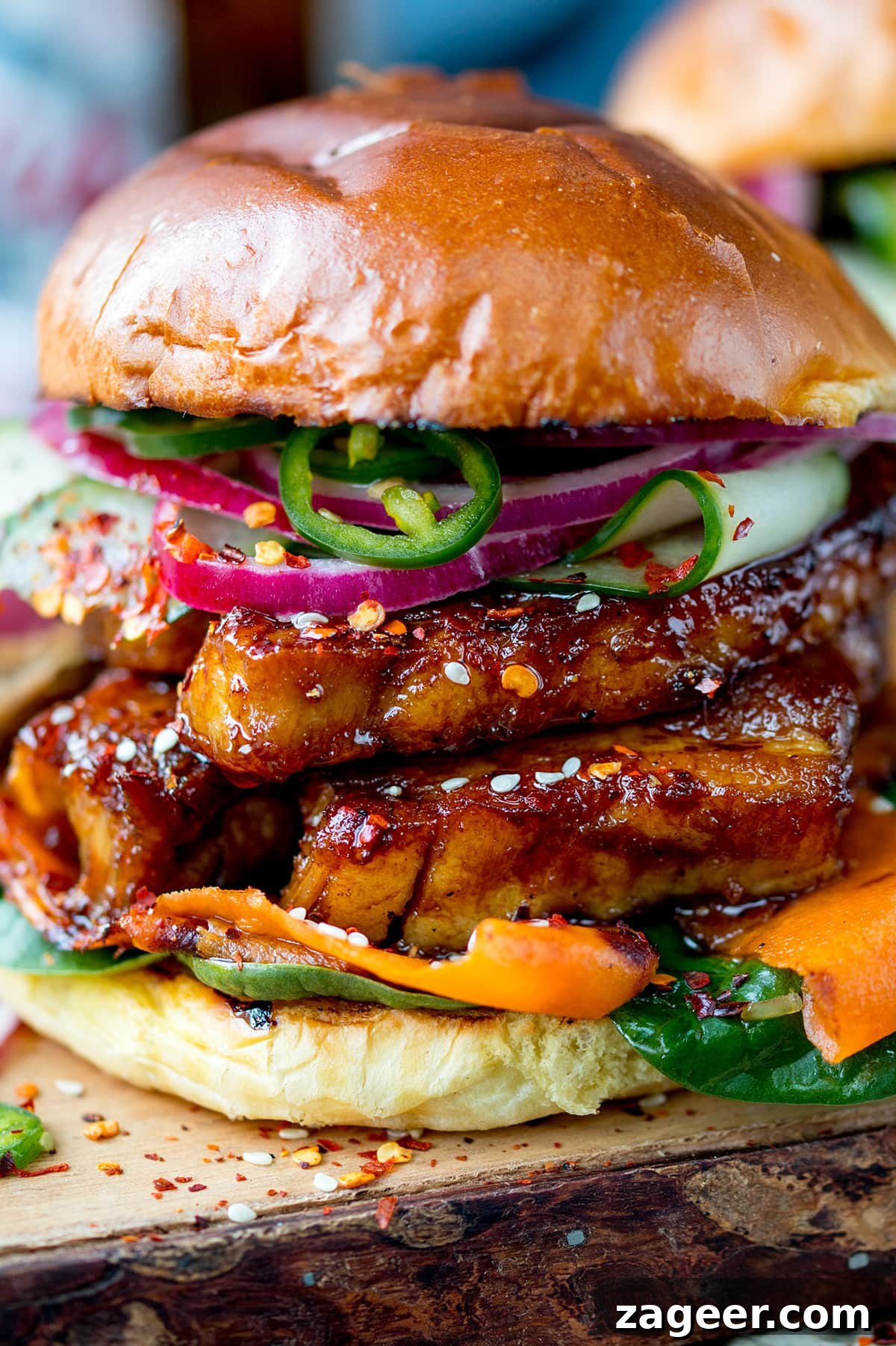 Sticky Glazed Pork Burger with Quick Pickled Veggies 6 Side on, very close up image of a sticky pork belly burger with carrot, spinach, red onion and jalapeños on a brioche bun. The burger is on a wooden board.