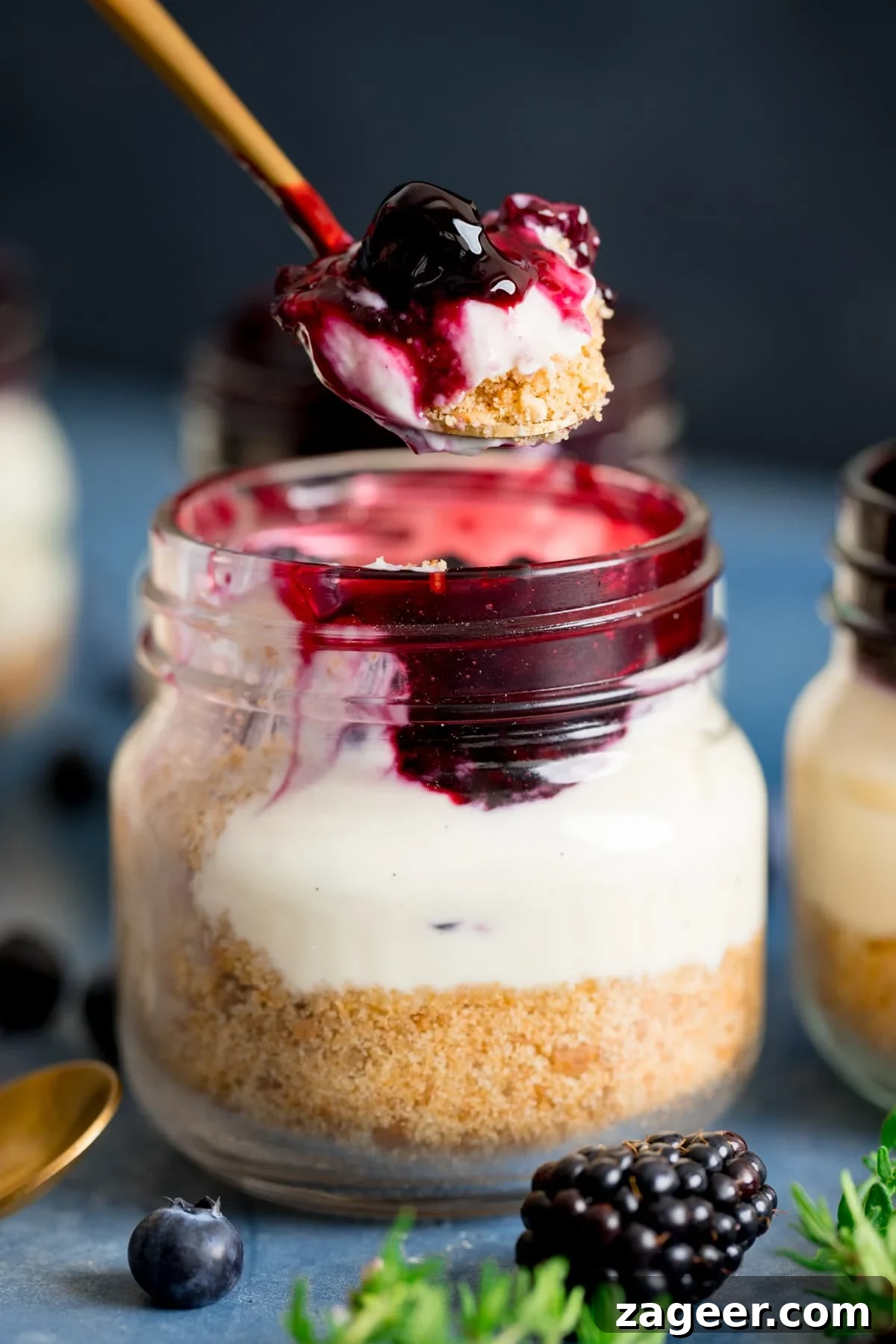 Mini No-Bake Berry Cheesecakes: Individual Dessert Cups 2 Spoonful being taken from a mason jar cheesecake on a blue background. Thyme leaves, blackberry and blueberries also in shot.