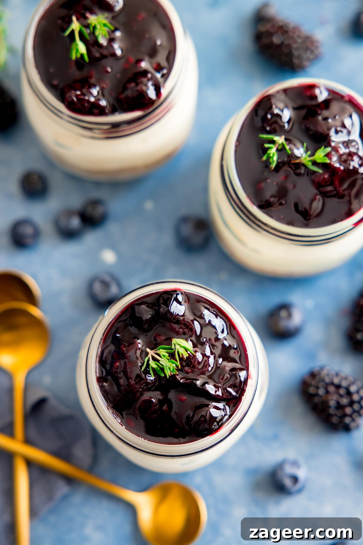 Mini No-Bake Berry Cheesecakes: Individual Dessert Cups 11 Overhead image of 3 mason jar individual berry cheesecakes on a blue background. Berries and gold teaspoons are next to the cheesecakes