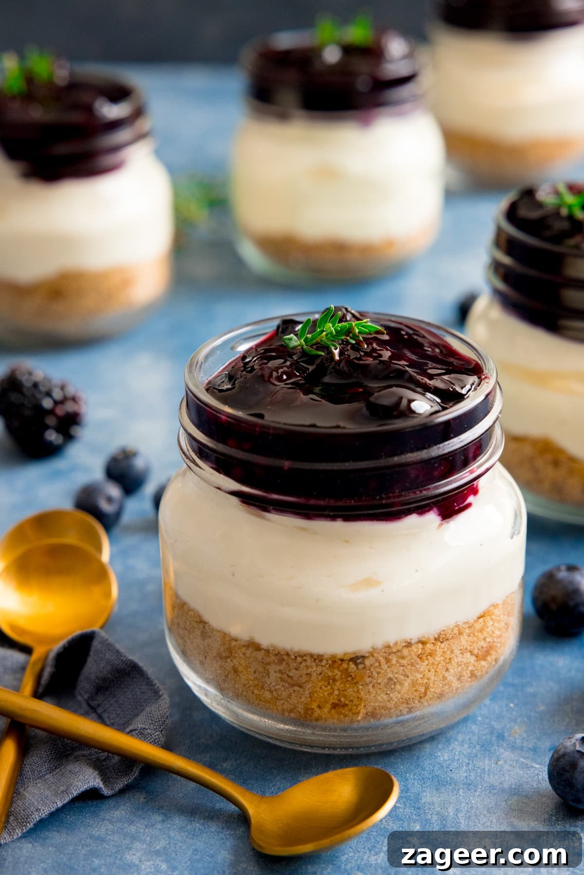 Mini No-Bake Berry Cheesecakes: Individual Dessert Cups 10 Mason jar berry cheesecake on a blue surface next to two gold teaspoons. Further mason jar cheesecakes in the background.
