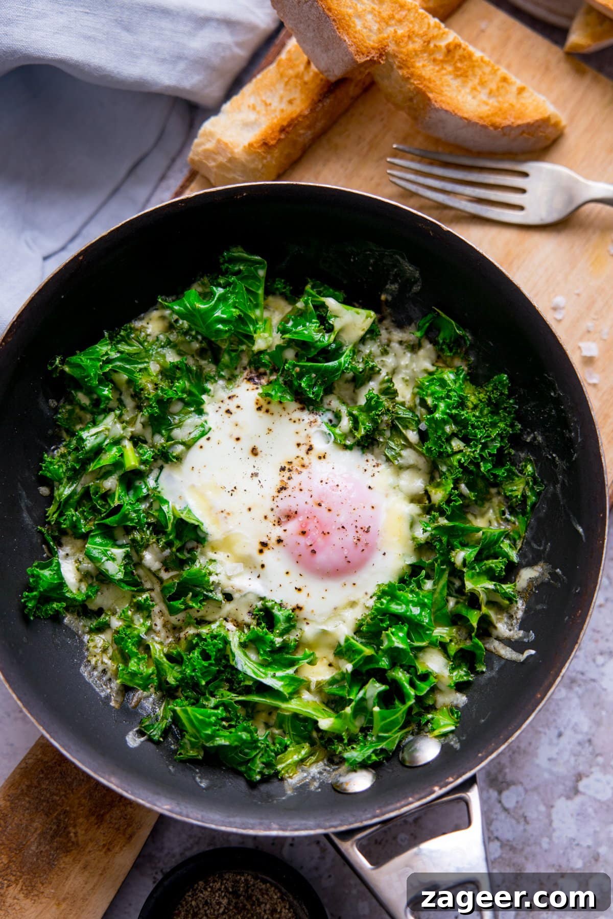 Creamy Scrambled Eggs with Sautéed Kale Breakfast Bowl 3 Fried egg in a pan with kale and cheese. Pan is on a wooden board with toast soldiers at the top of the shot.