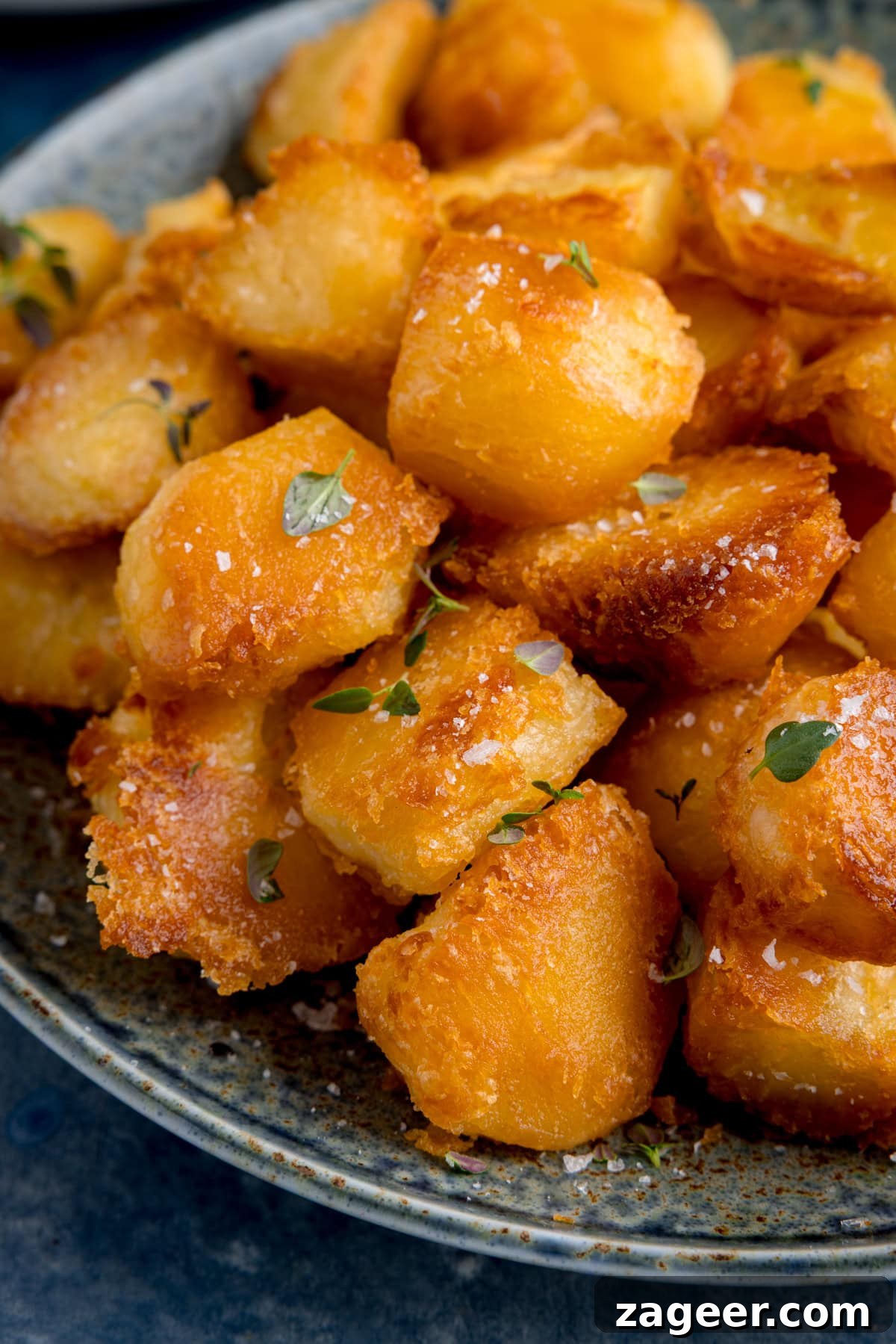 A close-up of golden, crispy perfect roast potatoes garnished with sea salt and fresh thyme on a speckled ceramic plate.