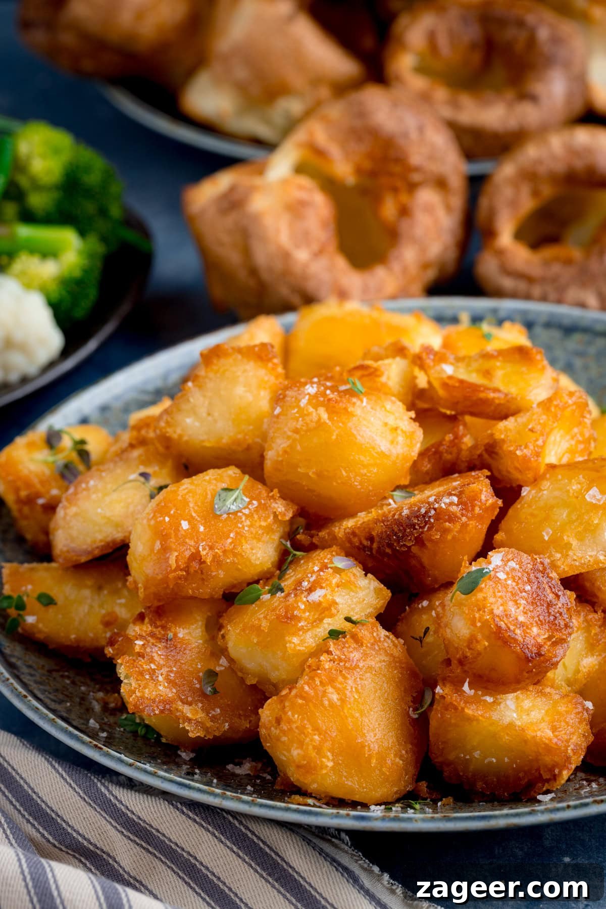 A plate of golden, crispy roast potatoes garnished with herbs, with Yorkshire puddings and vegetables visible in the background.