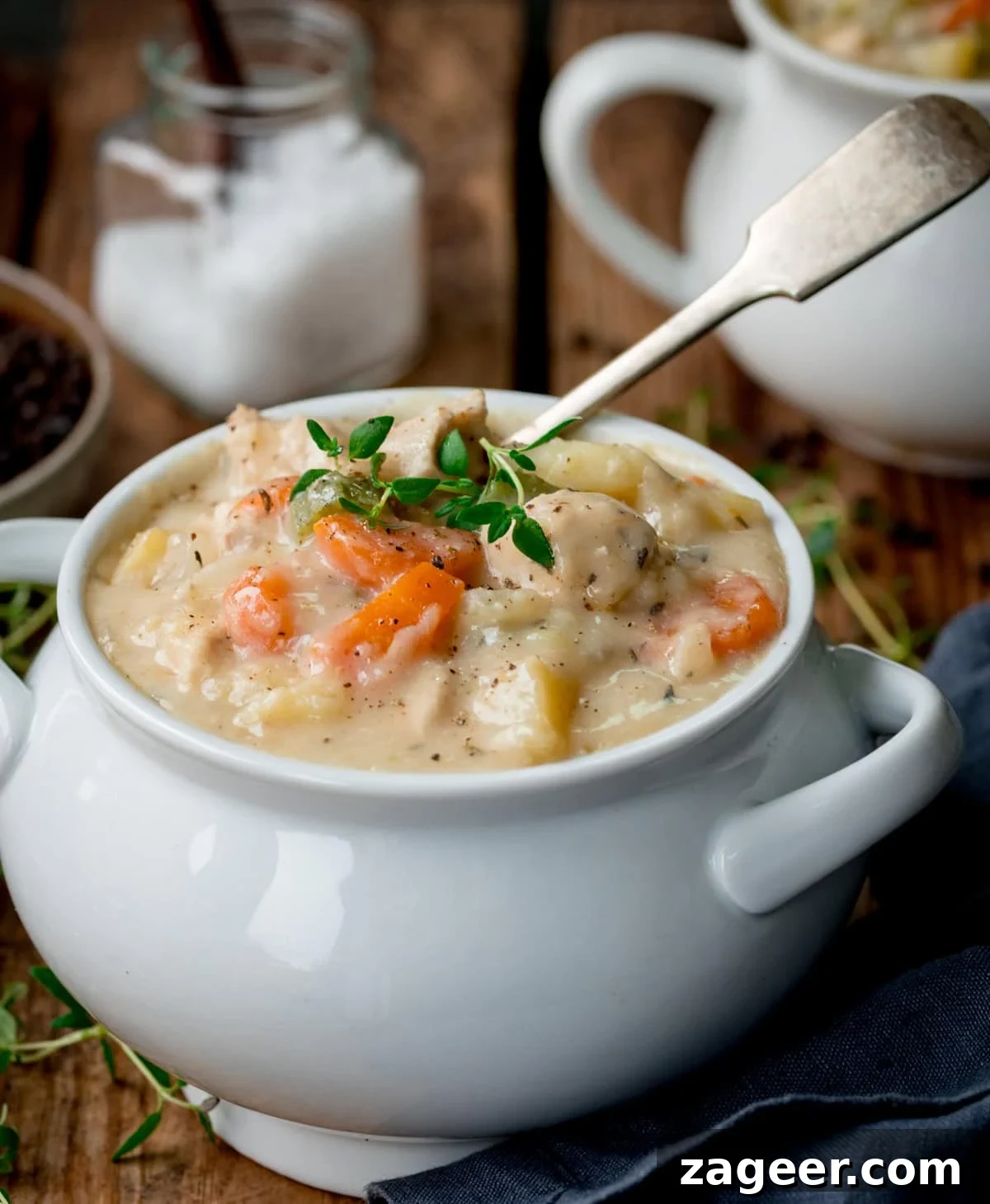 Creamy Chicken Pot Pie Soup Recipe — Comfort in a Bowl 4 A bowl of creamy chicken pot pie soup with vegetables, garnished with herbs, sits on a wooden table with a spoon in the bowl. A jar of salt and another bowl are in the background.