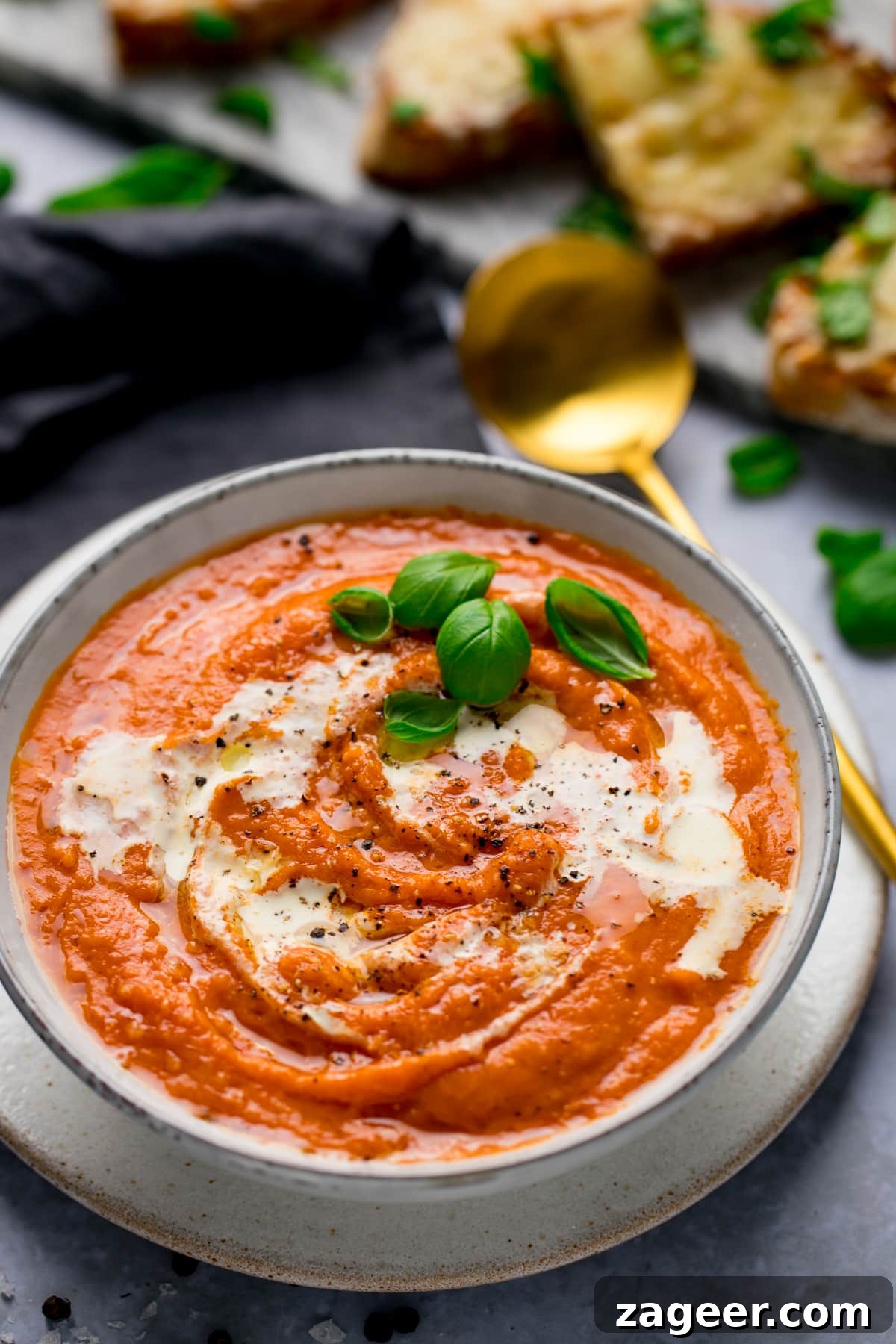 Homemade Creamy Tomato Soup Recipe from Scratch 6 Lush bowl of wholesome tomato soup covered with rich cream and basil leaves with a golden spoon. A slate of grilled cheese toast covered with basil leaves in the background.