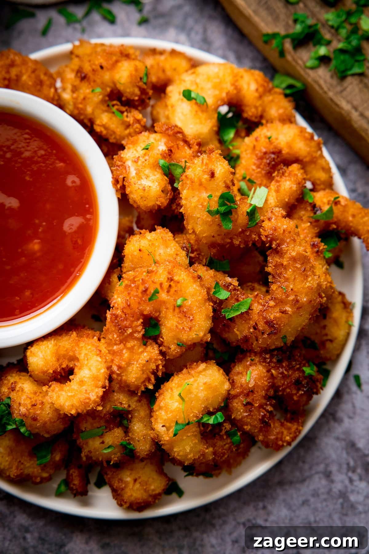 Crispy Coconut Shrimp Recipe with Mango Dipping Sauce 2 Coconut shrimp on a plate on a grey background. Dish of sweet and sour sauce is next to the prawns.