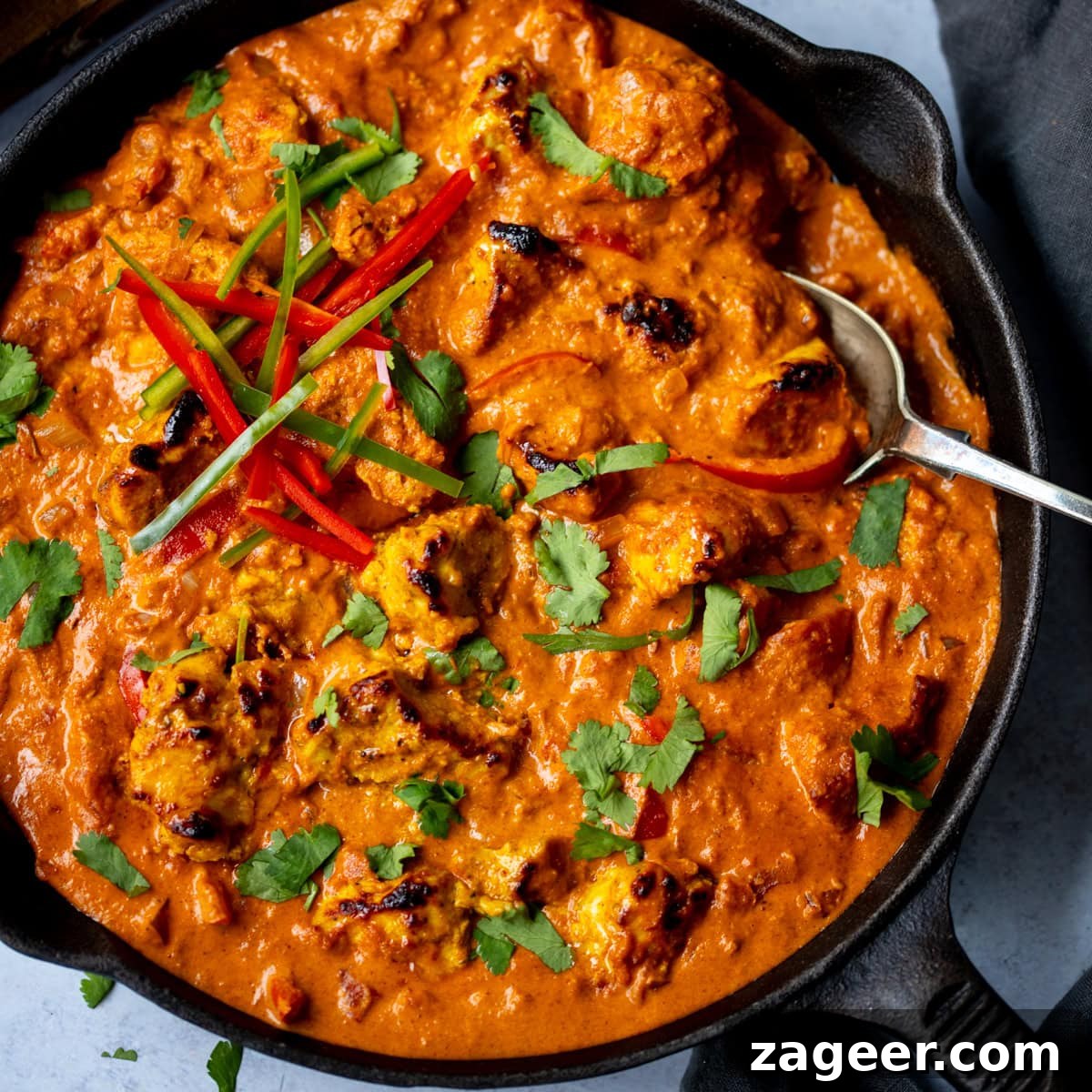Authentic Chicken Tikka Masala Recipe for Tender, Creamy Curry 2 A frying pan filled with Chicken Tikka Masala, garnished with coriander and sliced red chilli, with a spoon resting inside.