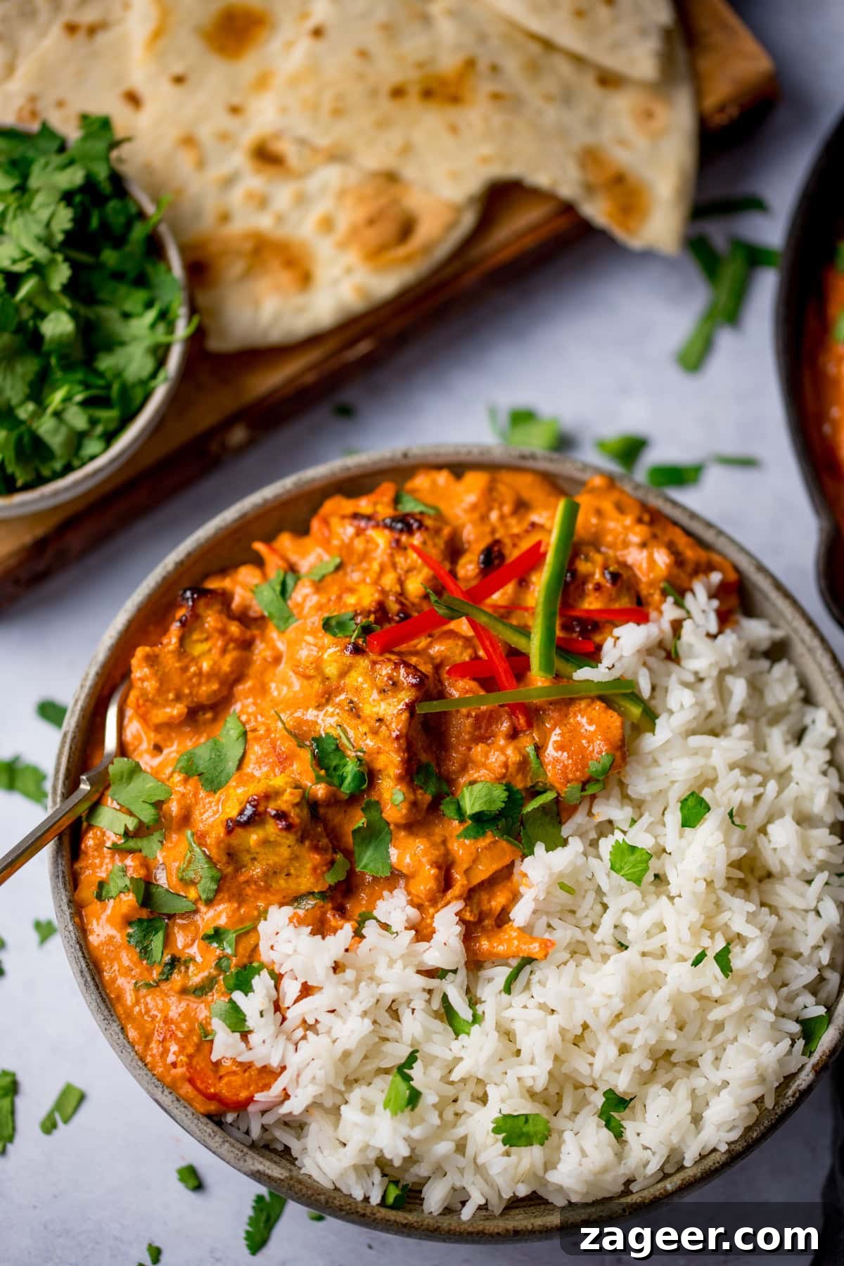 Authentic Chicken Tikka Masala Recipe for Tender, Creamy Curry 15 A bowl Chicken Tikka Masala and rice, garnished with coriander and red chilli, served with naan bread and fresh coriander on the side.