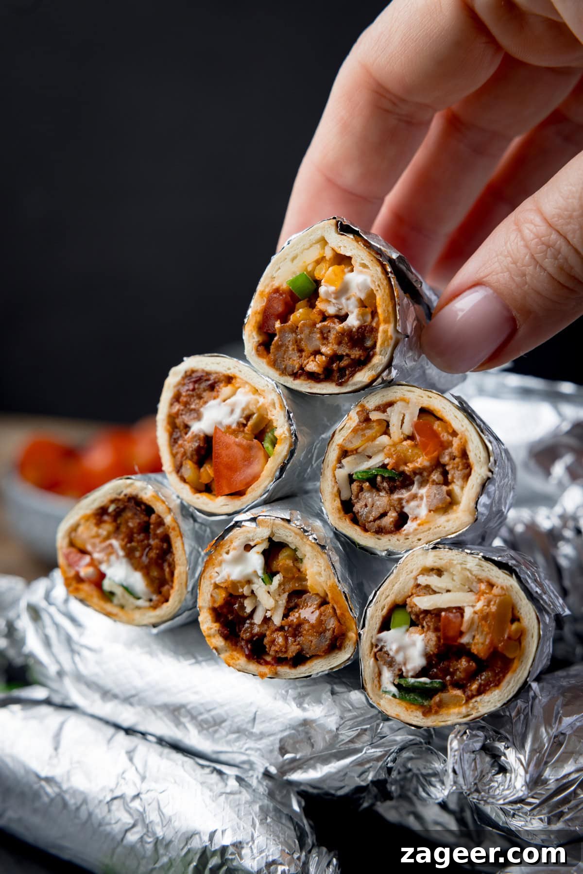 Mini Chicken and Bean Burritos with Zesty Salsa Recipe 2 Tall image showing mini burritos wrapped in foil and piled up with a black background. Some of the burritos have been sliced in half and piled on top of the whole burritos to show the filling. A hand is holding picking up one of the open burritos. There is a small bowl of tomatoes blurred out in the background.