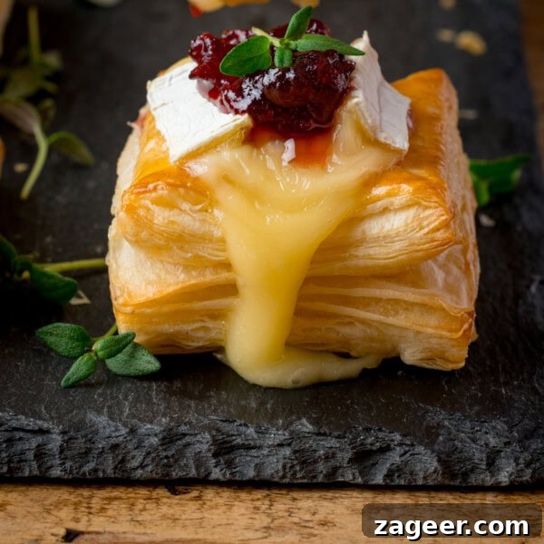 Mini Chicken and Bean Burritos with Zesty Salsa Recipe 14 Close up of a square puff pastry topped with melted brie cheese and cranberry sauce, garnished with fresh thyme, sitting on a dark slate serving board.