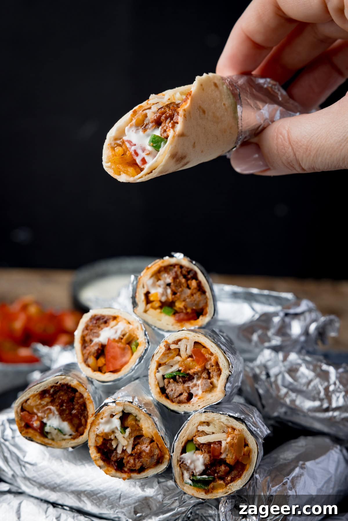Mini Chicken and Bean Burritos with Zesty Salsa Recipe 8 Tall image showing mini burritos wrapped in foil, piled up on a plate against a black background. Some of the burritos have been sliced in half and piled on top of the whole burritos to show the filling. A hand is holding an unwrapped burrito above the pile. There is a small bowl of tomatoes blurred out in the background.