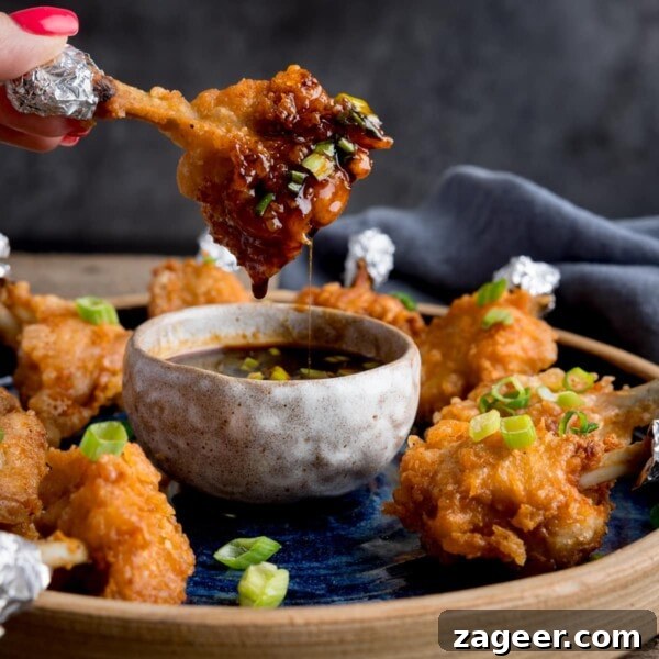 Mini Chicken and Bean Burritos with Zesty Salsa Recipe 9 Square image of a plate of chicken lollipops with a bowl of spicy dipping sauce. One of the wings is being dipped into the sauce and is dripping. The wing is being held by fingers with pink-painted fingernails.