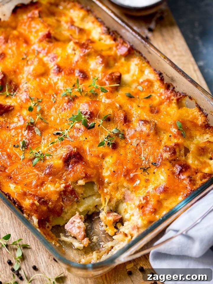Creamy Scalloped Potatoes with Ham and Cheese Bake 5 Scalloped potatoes with ham in a glass baking dish, scoop removed. The baking dish is sat on a wooden board with a light blue napkin in the foreground.