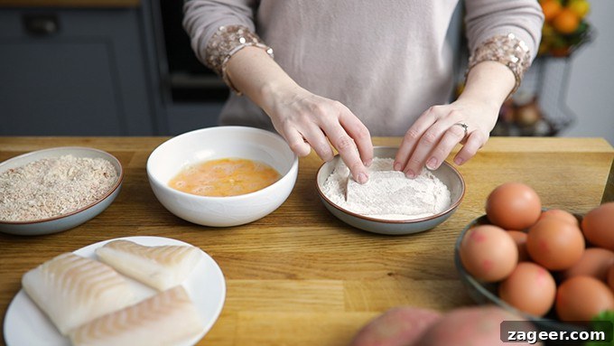 Crispy Fish Sandwich with Shoestring Fries Recipe 3 Hands dipping a haddock fillet into a bowl of flour. Bowl of whisked egg and a further bowl with breadcrumbs in scene. Plate of 3 haddock fillets and bowl of eggs partially in shot.