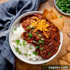 A bowl of chilli con carne with kidney beans, ground beef, cheese, and cilantro over white rice. Tortilla chips on the side.