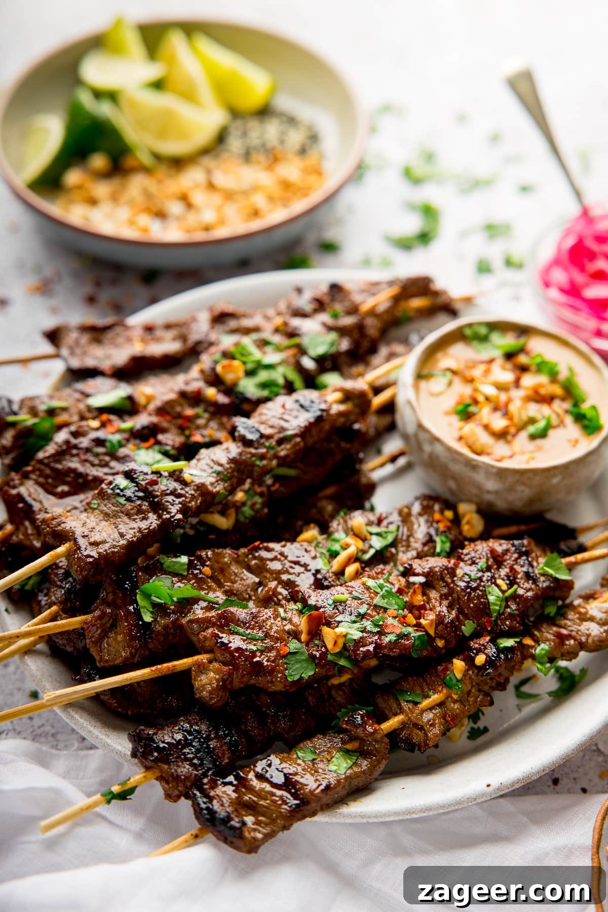 Grilled Beef Satay Recipe: Tender Skewers with Peanut Sauce 10 beef satay on a white plate with satay sauce. Bowl of chopped peanuts and lime in background.