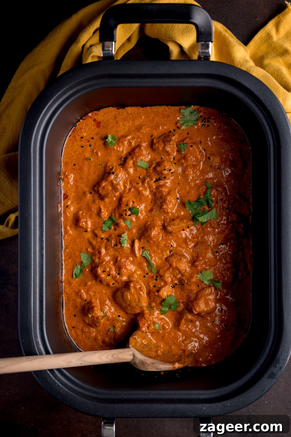 Slow-Cooker Butter Chicken Recipe for Tender, Creamy Meals 4 Overhead picture of crock pot butter chicken in the slow cooker bowl, sprinkled with a little fresh coriander with a wooden spoon in the corner.