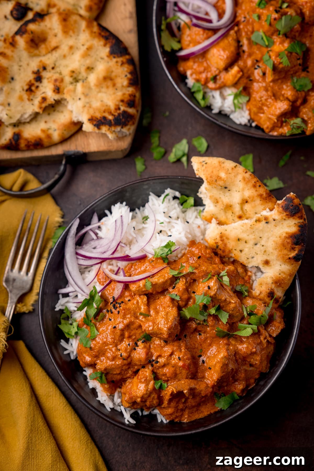 Slow-Cooker Butter Chicken Recipe for Tender, Creamy Meals 5 Overhead picture of a dark bowl full of boiled rice and crock pot butter chicken with some sliced red onion and charred naan bread in the corner of the bowl, with a fork and a board with more naan bread in the background.