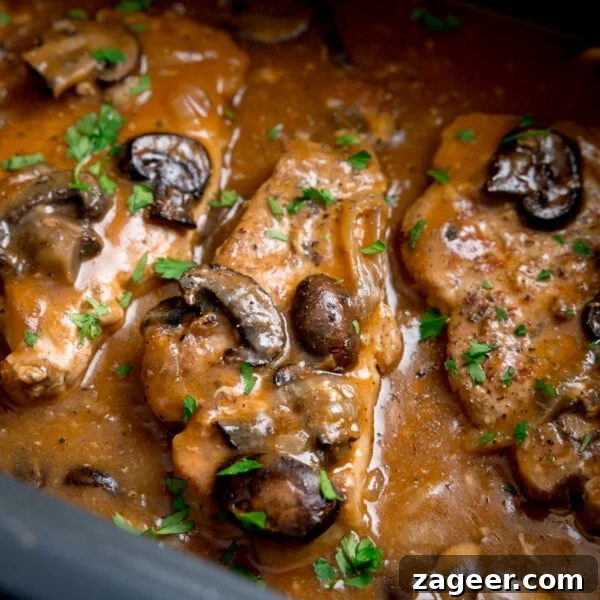 Slow-Cooker Butter Chicken Recipe for Tender, Creamy Meals 7 Square close-up image of smothered pork chops with mushrooms and gravy in the pan of a slow cooker. There is chopped parsley sprinkled on top.