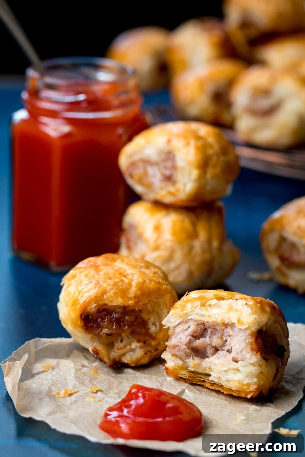 Crispy Homemade Sausage Rolls Recipe for Perfect Party Snacks 8 Sausage rolls and a blob of ketchup on a piece of parchment. Bite taken out of one of the sausage rolls.