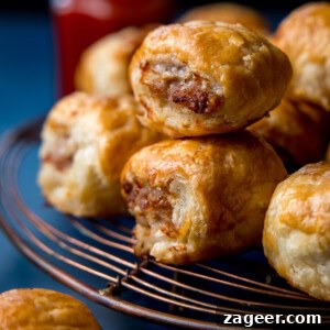 Crispy Homemade Sausage Rolls Recipe for Perfect Party Snacks 9 Several golden-brown sausage rolls stacked on a wire cooling rack, with a blurred bottle of sauce in the background.