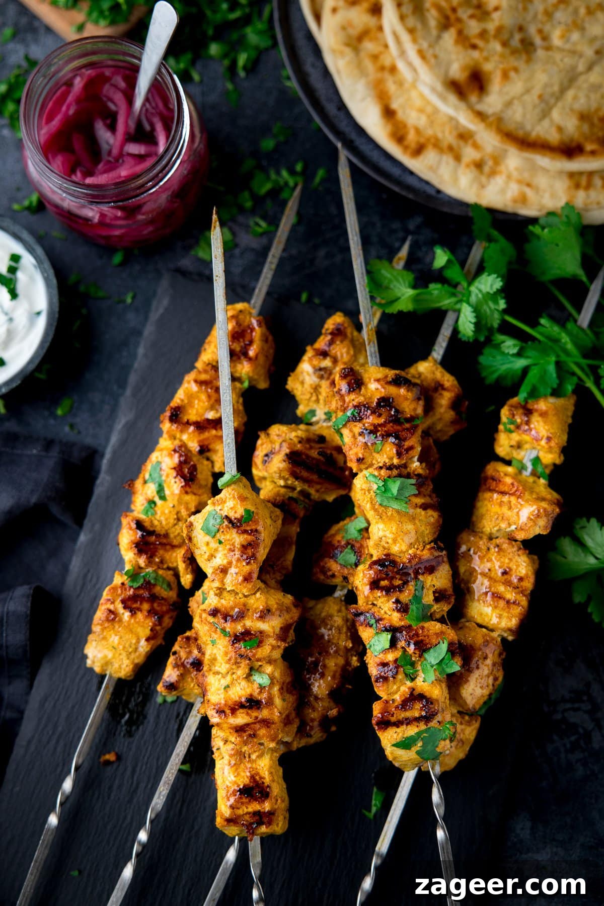 chicken tikka skewers on a dark background with pita bread and a jar of pickled red onion slices
