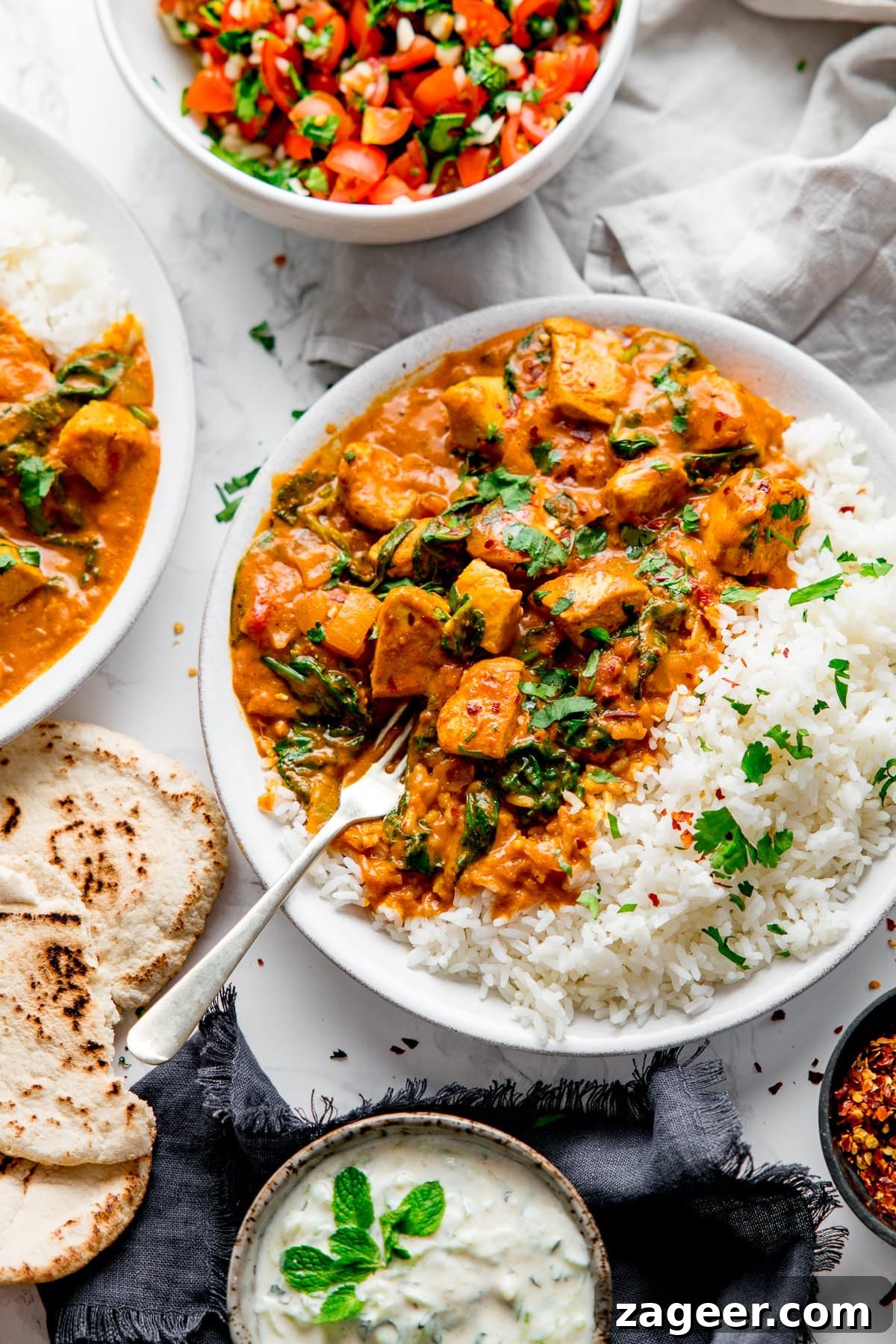 Simple Indian-Style Chicken Curry Recipe for Weeknight Dinners 6 White bowl with chicken curry and rice with a fork digging into the curry. Ingredients and side dishes scattered around.