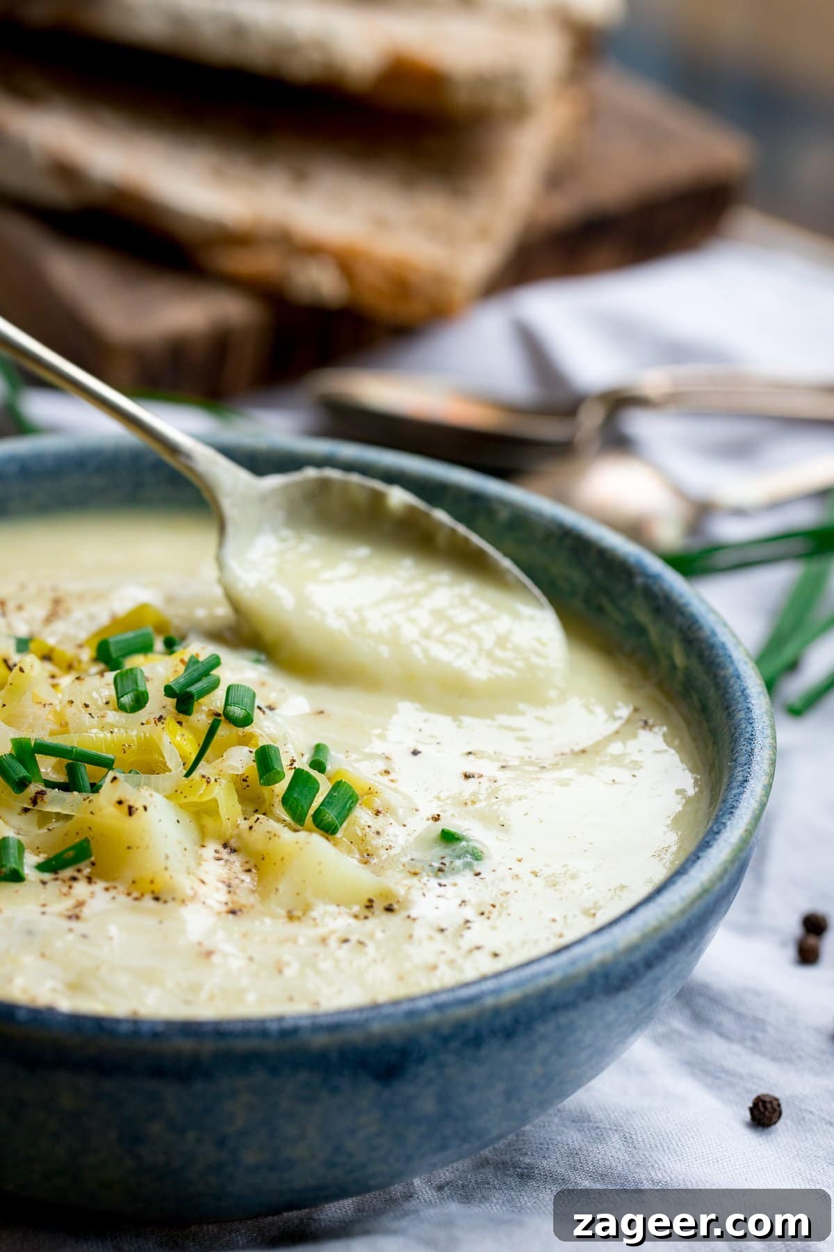 Creamy Leek and Potato Soup Recipe with Fresh Herbs 2 spoonful being taken from a bowl of leek and potato soup. Bowl is blue. Sliced bread in the background.