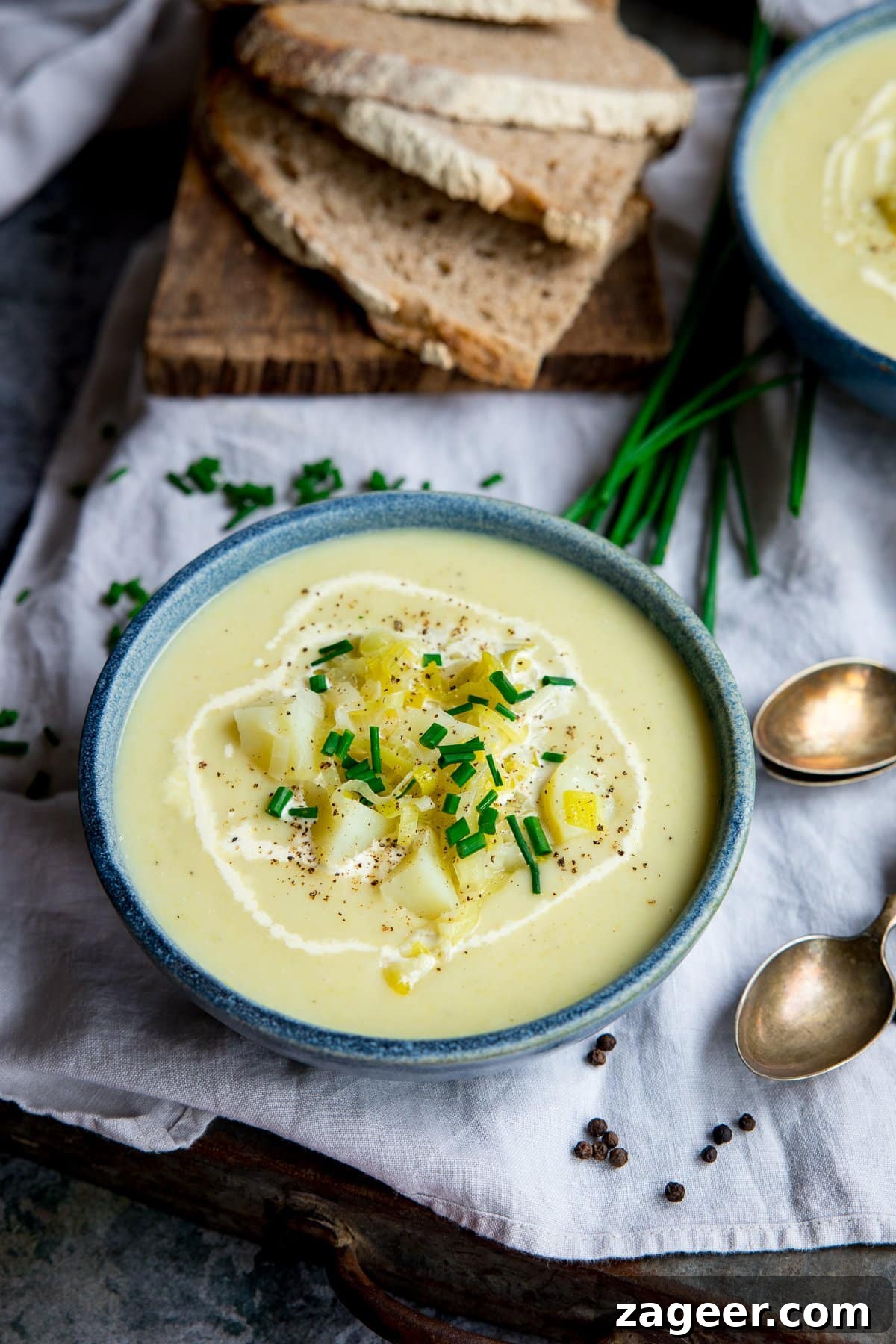 Creamy Leek and Potato Soup Recipe with Fresh Herbs 7 leek and potato soup in a blue bowl on top of a napkin. Sliced bread and further bowl at the top of the shot.