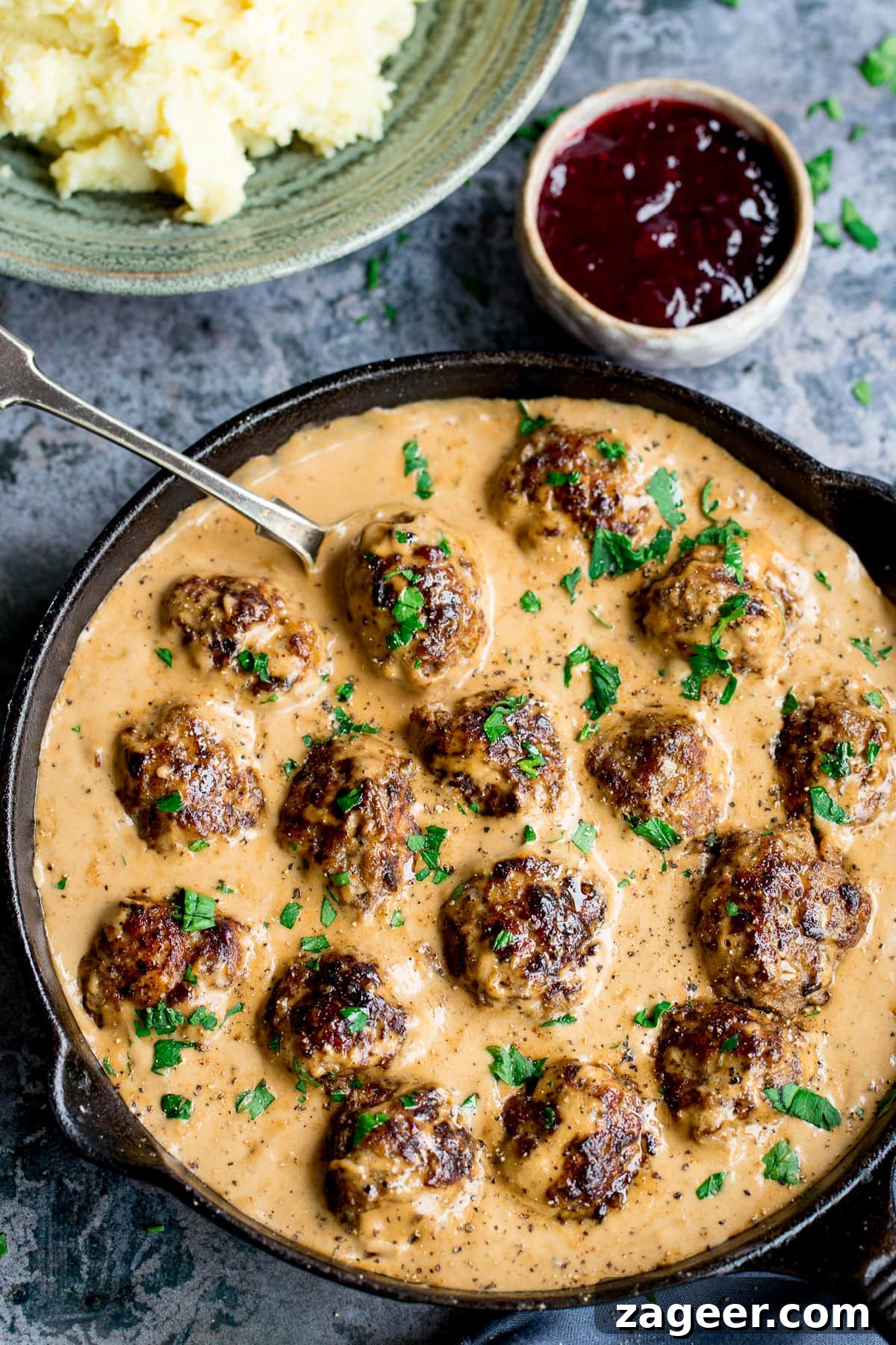 Authentic Swedish Meatball Recipe with Creamy Gravy and Lingonberry 7 Swedish meatballs and gravy in a pan next to a pot of lingonberry jelly and mashed potatoes