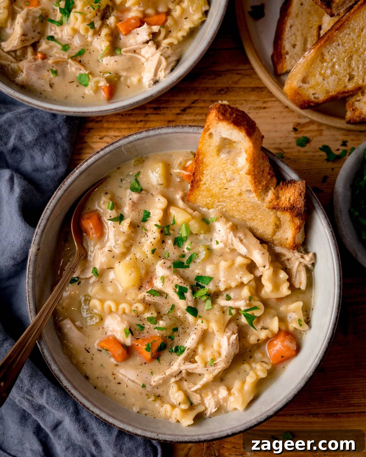 Classic Homemade Chicken Noodle Soup Recipe for Comforting Meals 2 A bowl of homemade chicken noodle soup with pasta, carrots, and potatoes, garnished with parsley and served with toasted buttered bread. The soup bowl is on a wooden table with another bowl just in shot at the top of the frame.