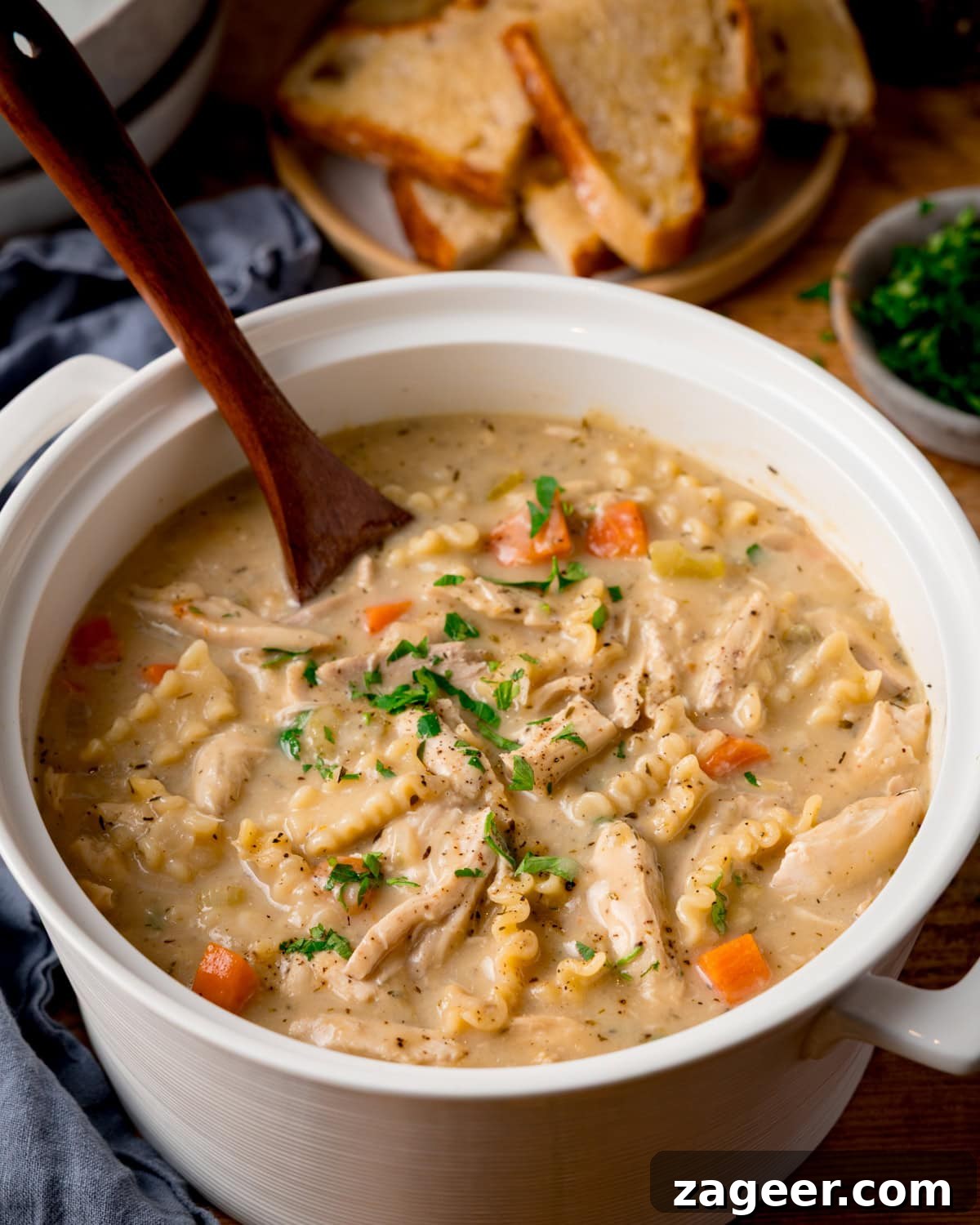 Classic Homemade Chicken Noodle Soup Recipe for Comforting Meals 5 A pot of creamy chicken noodle soup with carrots and herbs. There is a wooden spoon sticking out. A plate of buttered toast pieces is in the background alongside a small bowl of chopped parsley and a blue napkin.