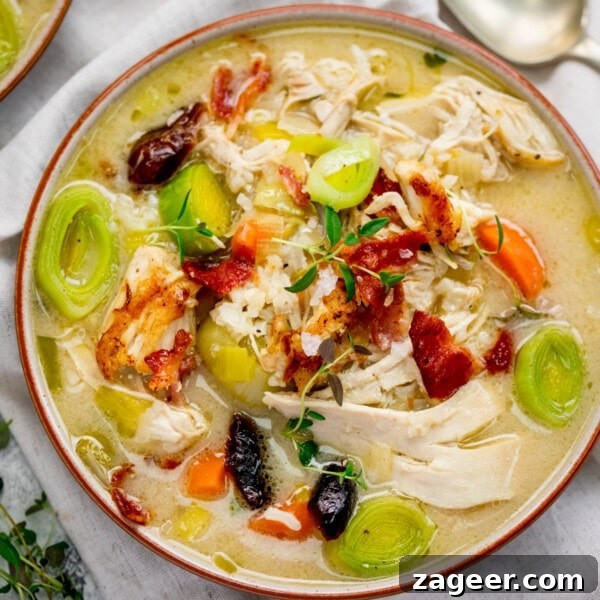 Classic Homemade Chicken Noodle Soup Recipe for Comforting Meals 7 A bowl of cock-a-leekie soup with sliced leeks, carrots, shredded chicken, bacon pieces, herbs and black olives sits on a white cloth with a spoon nearby.
