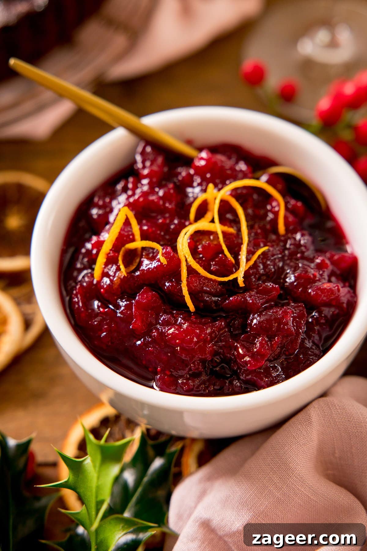 Port-Infused Cranberry Sauce Recipe for Holiday Menus 6 Cranberry sauce topped with orange zest in a white bowl. Festive decorations scattered around.