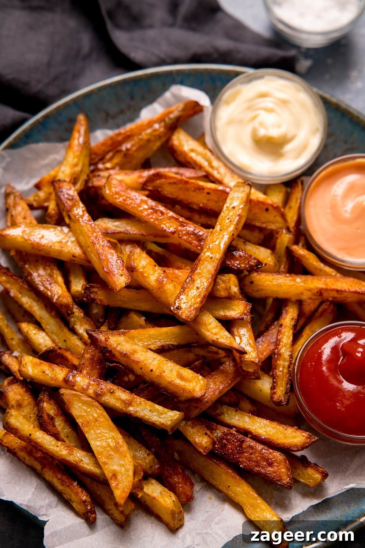 Crispy Homemade Oven Chips Recipe: Perfectly Golden Fries 2 Crispy oven chips on parchment paper on a blue-grey plate. Three bowls of dips on the plate.