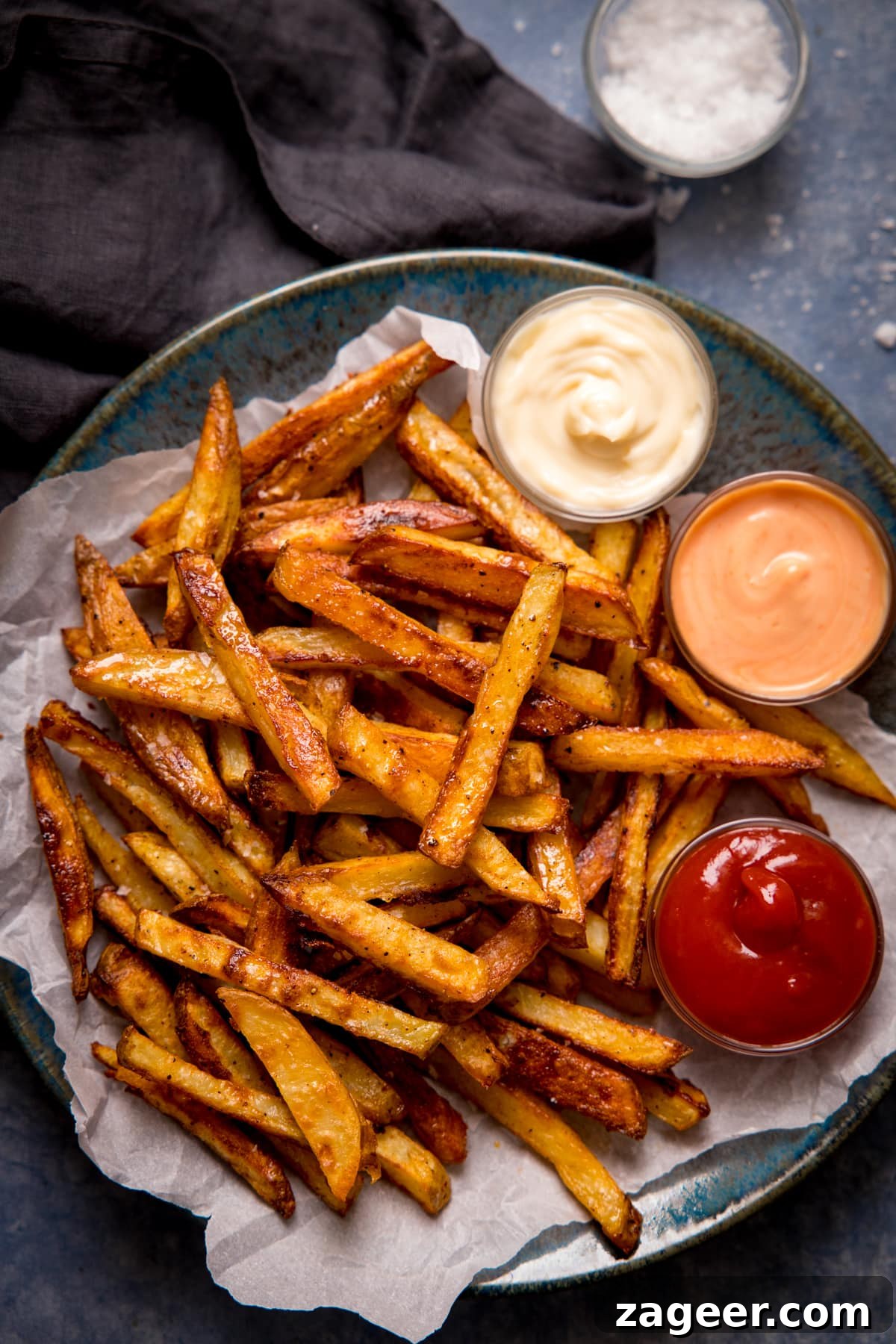 Crispy Homemade Oven Chips Recipe: Perfectly Golden Fries 6 Crispy oven fries and 3 dips on a plate on a grey-blue background.