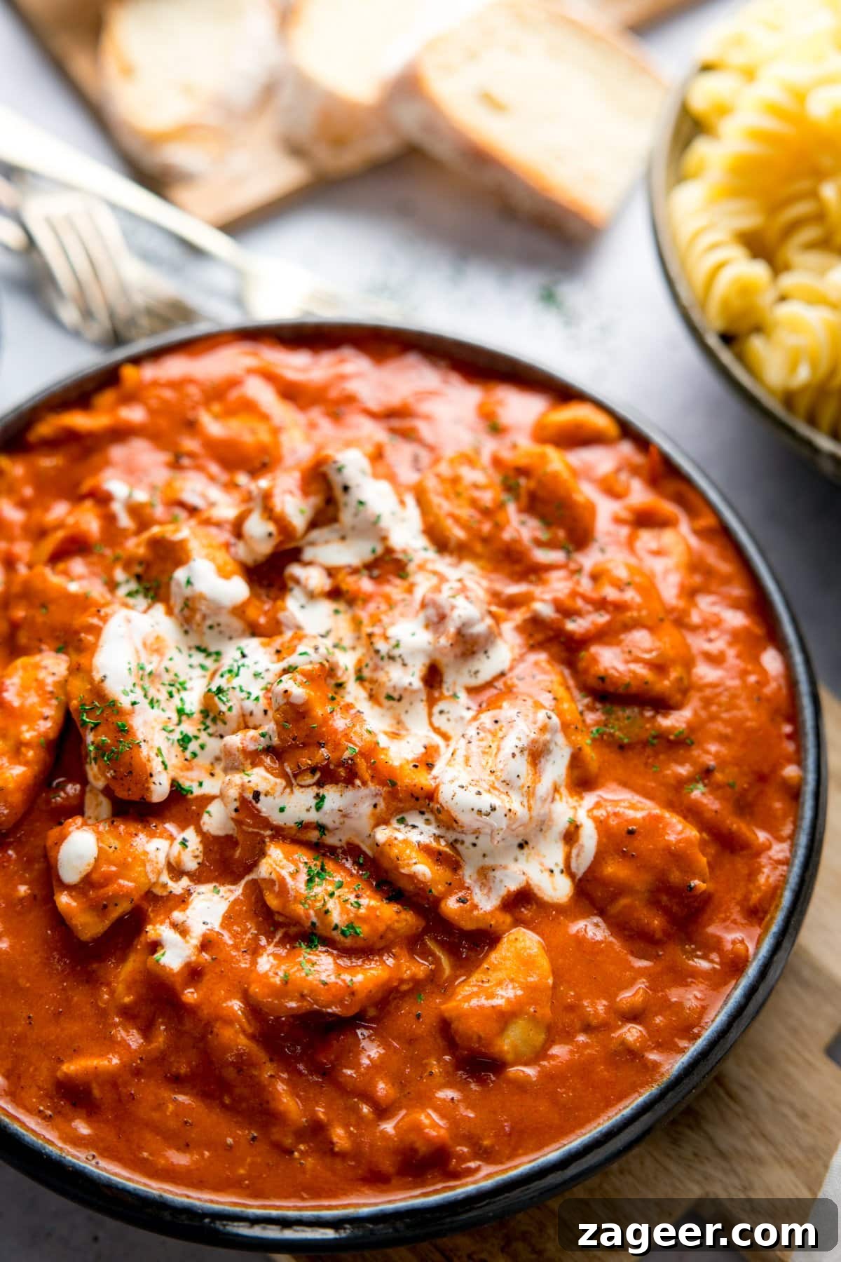Classic Hungarian Chicken Paprikash Recipe — Creamy Paprika Chicken 6 Dark bowl filled with chicken paprikash and topped with a swirl of soured cream. Bowl is on a light background and there are side dishes of bread and pasta just in shot.