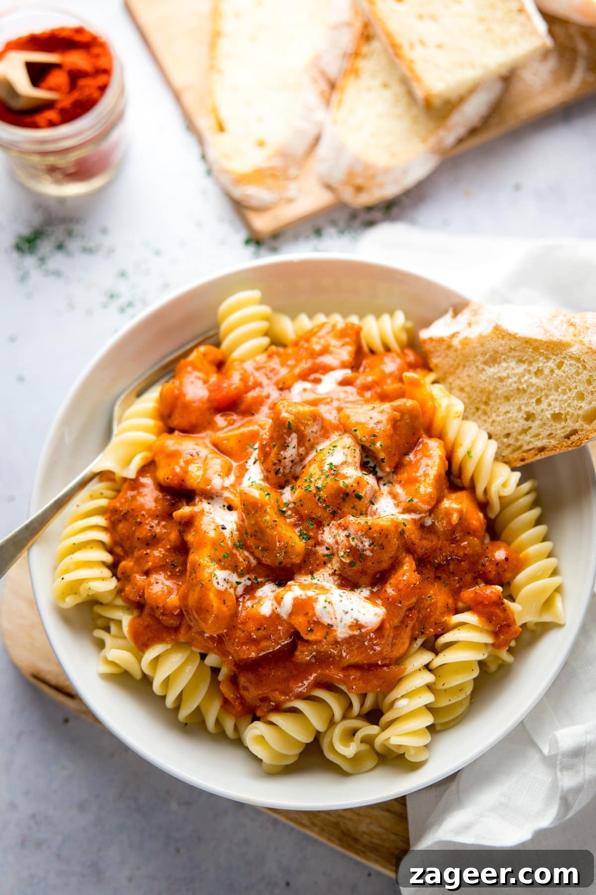 Classic Hungarian Chicken Paprikash Recipe — Creamy Paprika Chicken 7 Overhead image of a white bowl filled with chicken paprikash, pasta and a piece of bread. Bowl is on a light background and there is more bread a napkin and jar of paprika in shot.