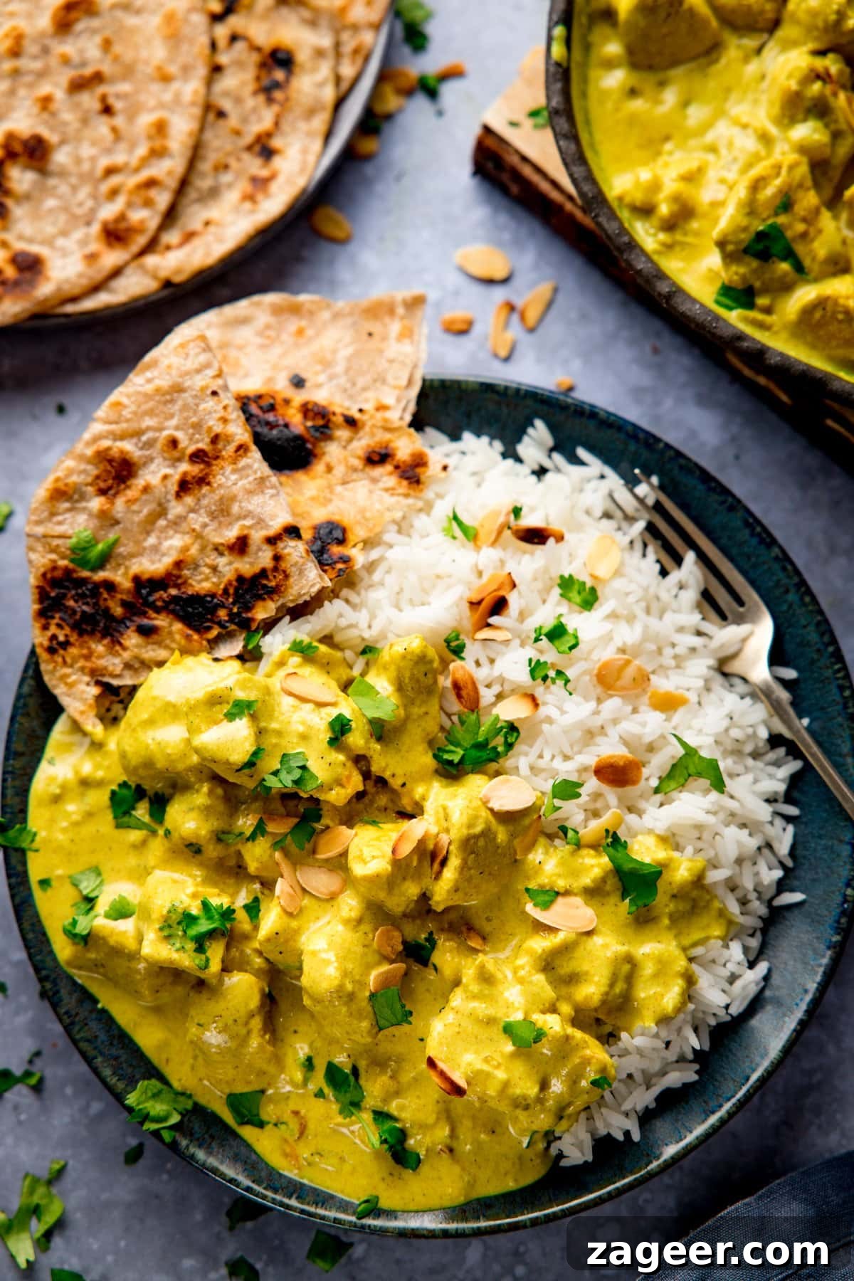 Creamy Chicken Pasanda Recipe: Aromatic Mughlai Curry Guide 2 Dark plate with rice, chapati and chicken pasanda - topped with almonds and coriander. All on light blue background with further dishes just in shot at the top of the frame.
