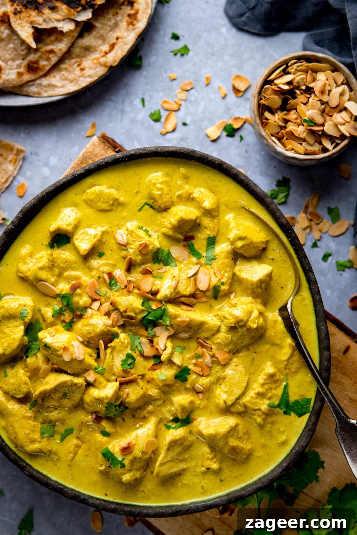 Creamy Chicken Pasanda Recipe: Aromatic Mughlai Curry Guide 12 Dark bowl filled with Chicken pasanda. Bowl is sitting on a wooden chopping board on a light blue background. Further ingredients scattered around.